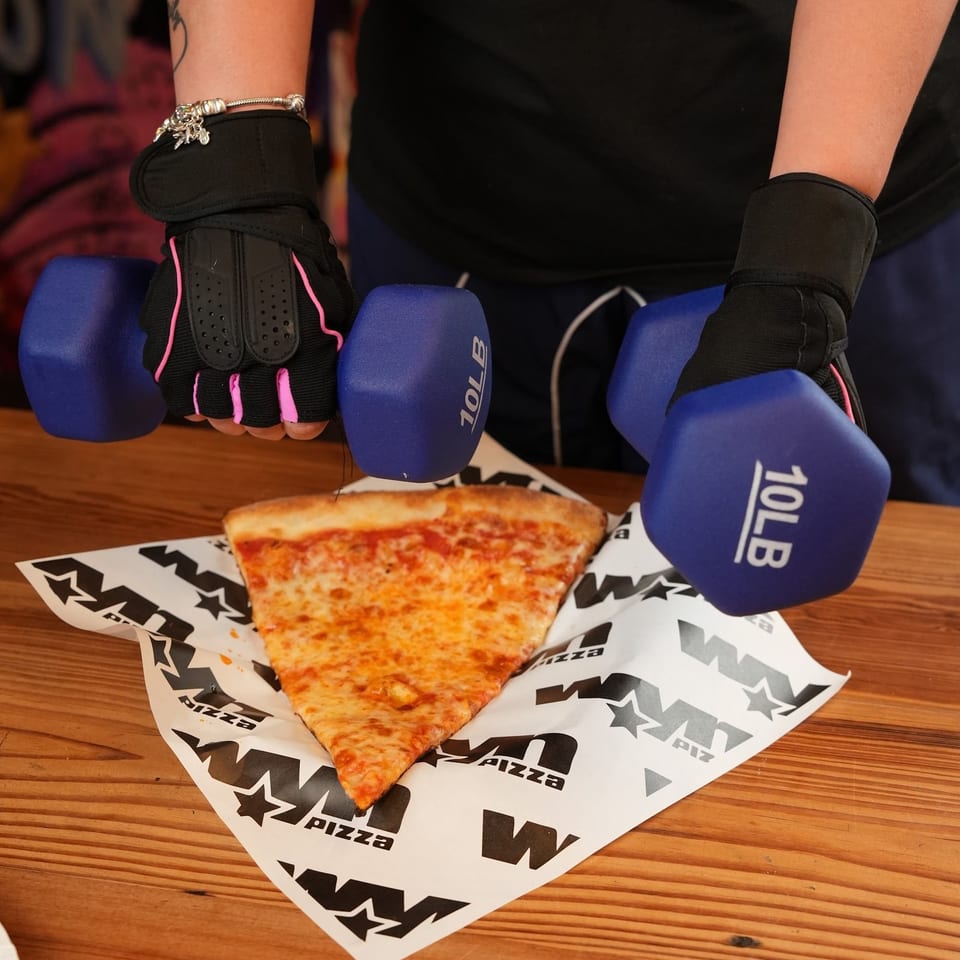 Person wearing black workout gloves holds blue dumbbells above a cheese pizza slice on branded paper.