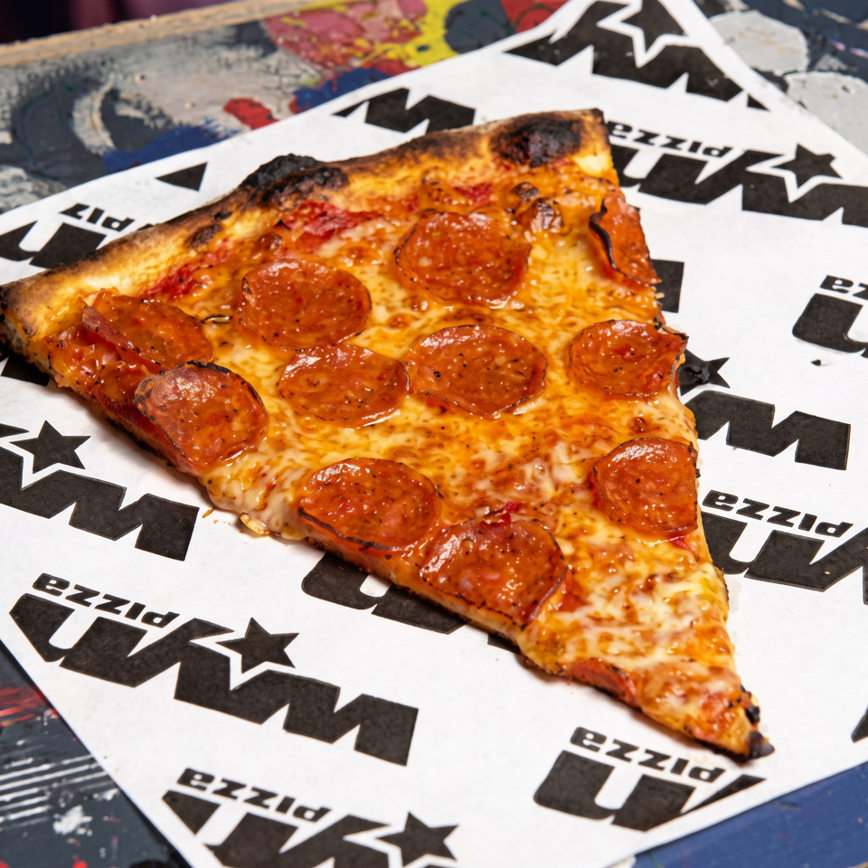 Pepperoni pizza slice with melted cheese and a charred crust served on branded paper.
