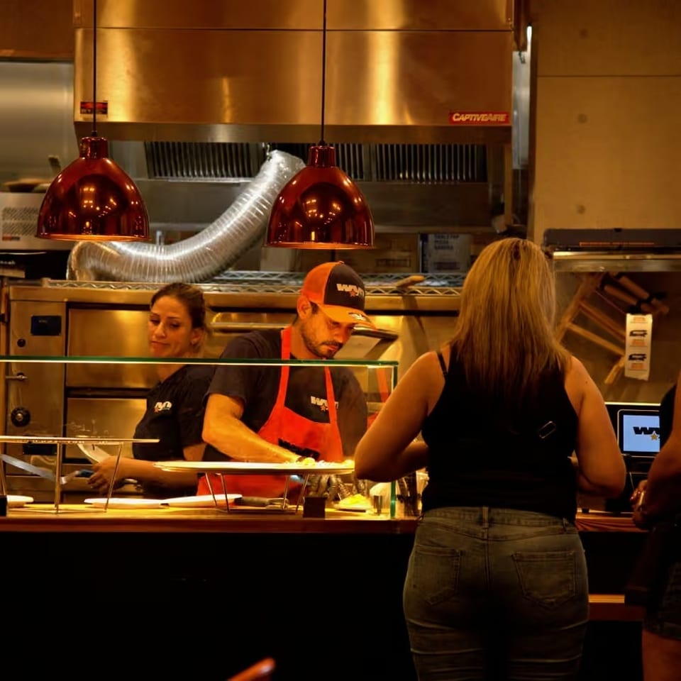 Restaurant counter with staff preparing food behind glass, red pendant lights, and a customer ordering at the open kitchen.